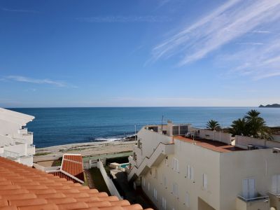 Vista desde la azotea con tejado de terracota, ofreciendo una panorámica del mar Mediterráneo y la playa de guijarros. Edificios costeros blancos y un cabo lejano bajo un cielo azul claro.