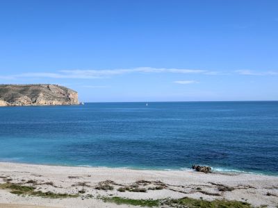 Vista panorámica de una playa de guijarros con aguas turquesas del Mediterráneo, un velero solitario en el horizonte y un acantilado rocoso a la izquierda.