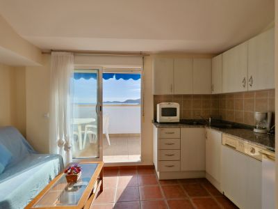 Zona de estar diáfana con cocina compacta de armarios blancos y suelo de baldosas de terracota. Puerta corredera de cristal a un balcón con vistas al mar y toldo azul.
