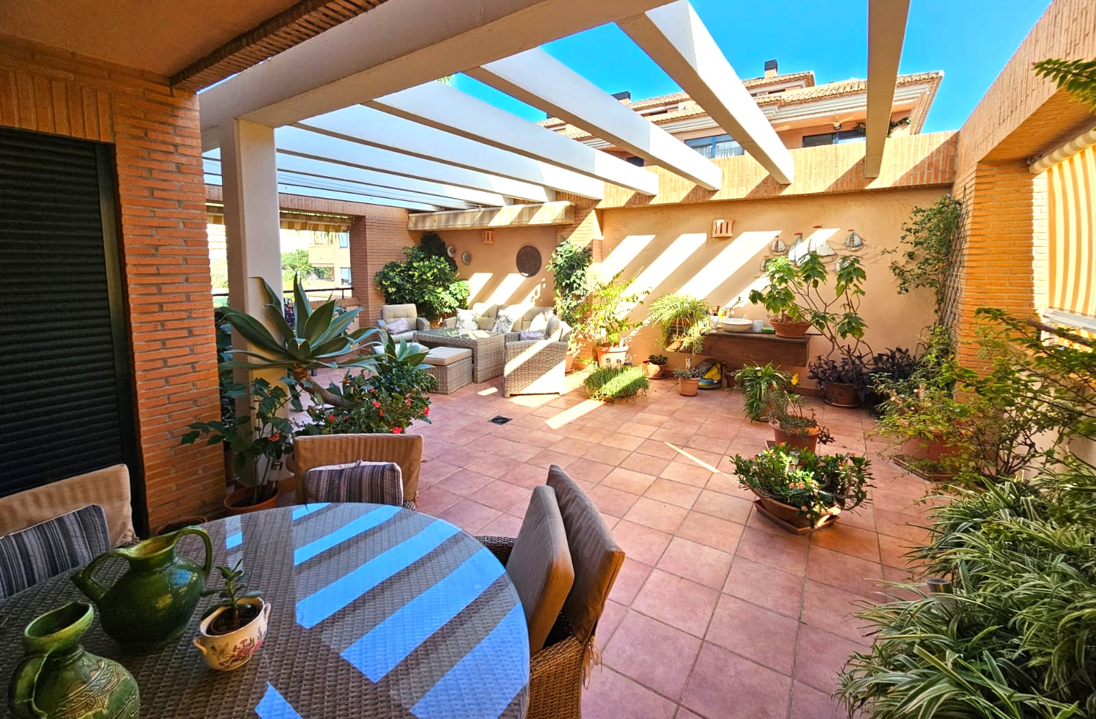 Amplia terraza mediterránea con suelo de barro, pérgola de vigas blancas y zonas de comedor y descanso rodeadas de abundante vegetación en macetas.