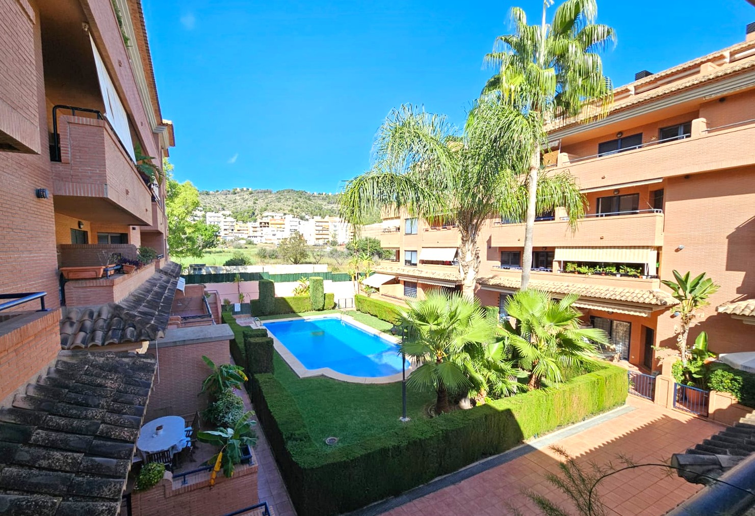 Vista desde el balcón a la zona comunitaria con piscina, césped cuidado y palmeras, rodeada de edificios residenciales de ladrillo con montañas al fondo.
