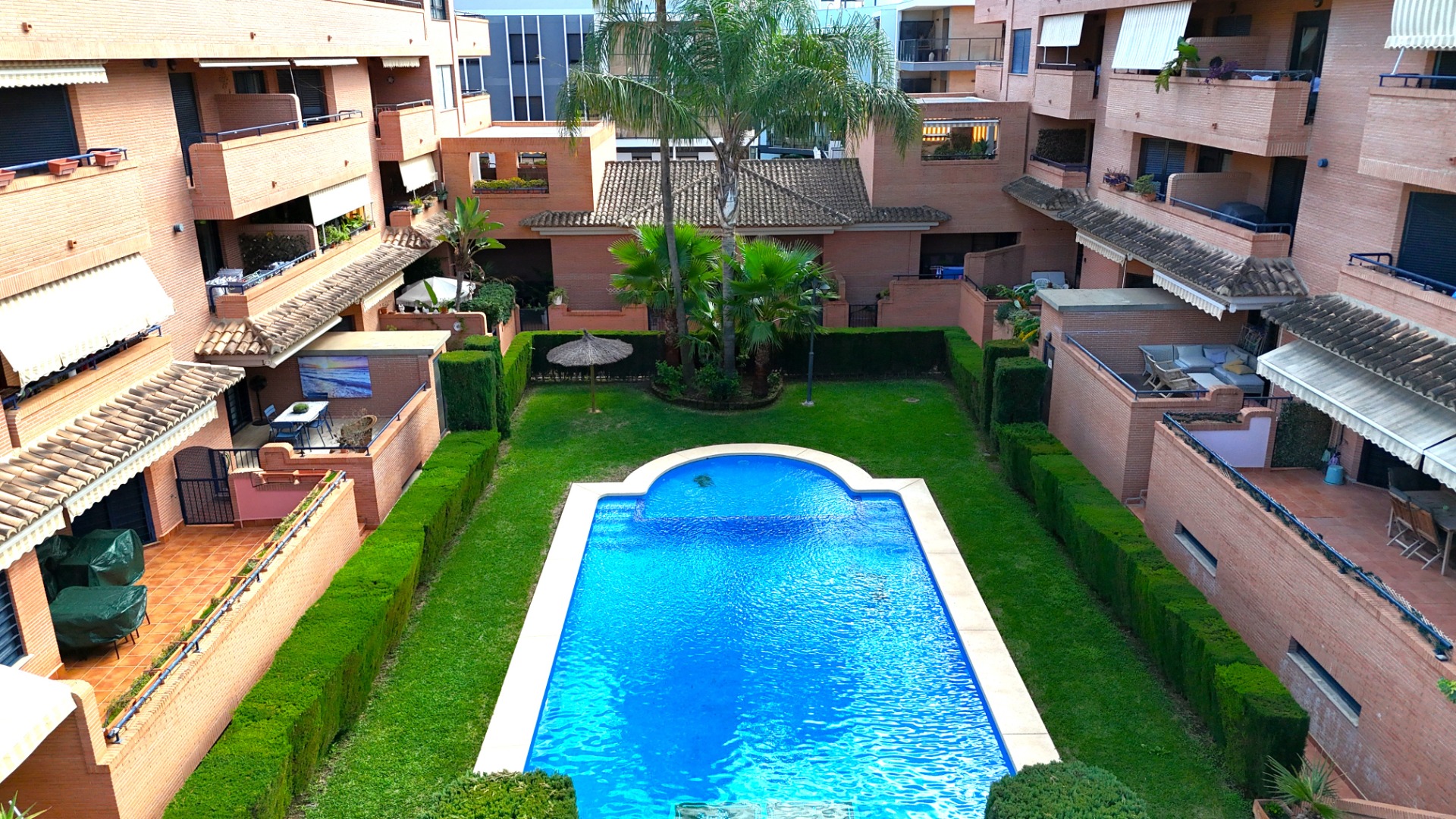 Zona comunitaria con piscina rectangular rodeada de césped, setos cuidados y edificios de ladrillo visto con amplias terrazas privadas y toldos.