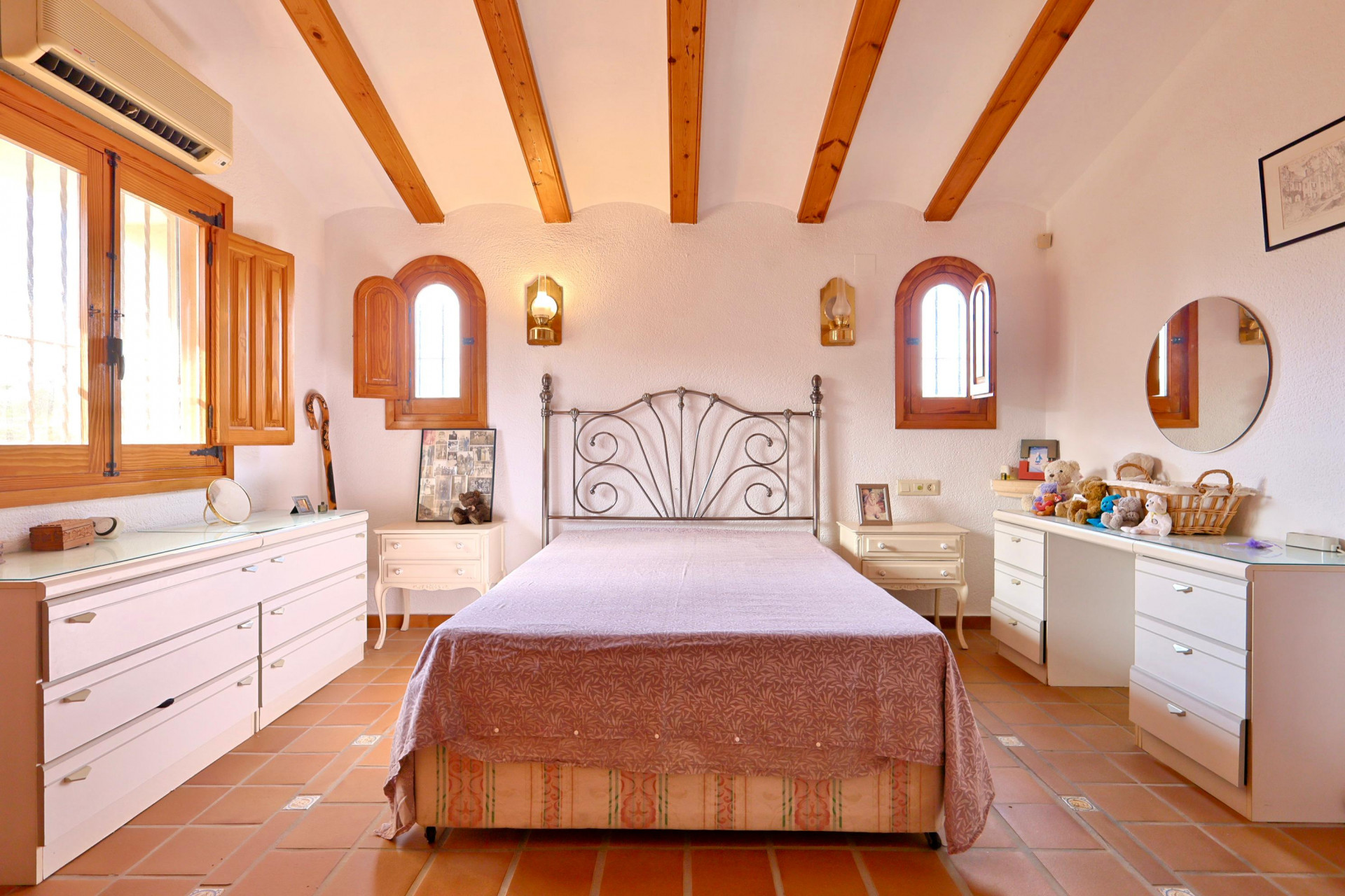 Dormitorio con vigas de madera a la vista, suelos de terracota y ventanas de arco con contraventanas de madera. Incluye cama de forja y aire acondicionado.