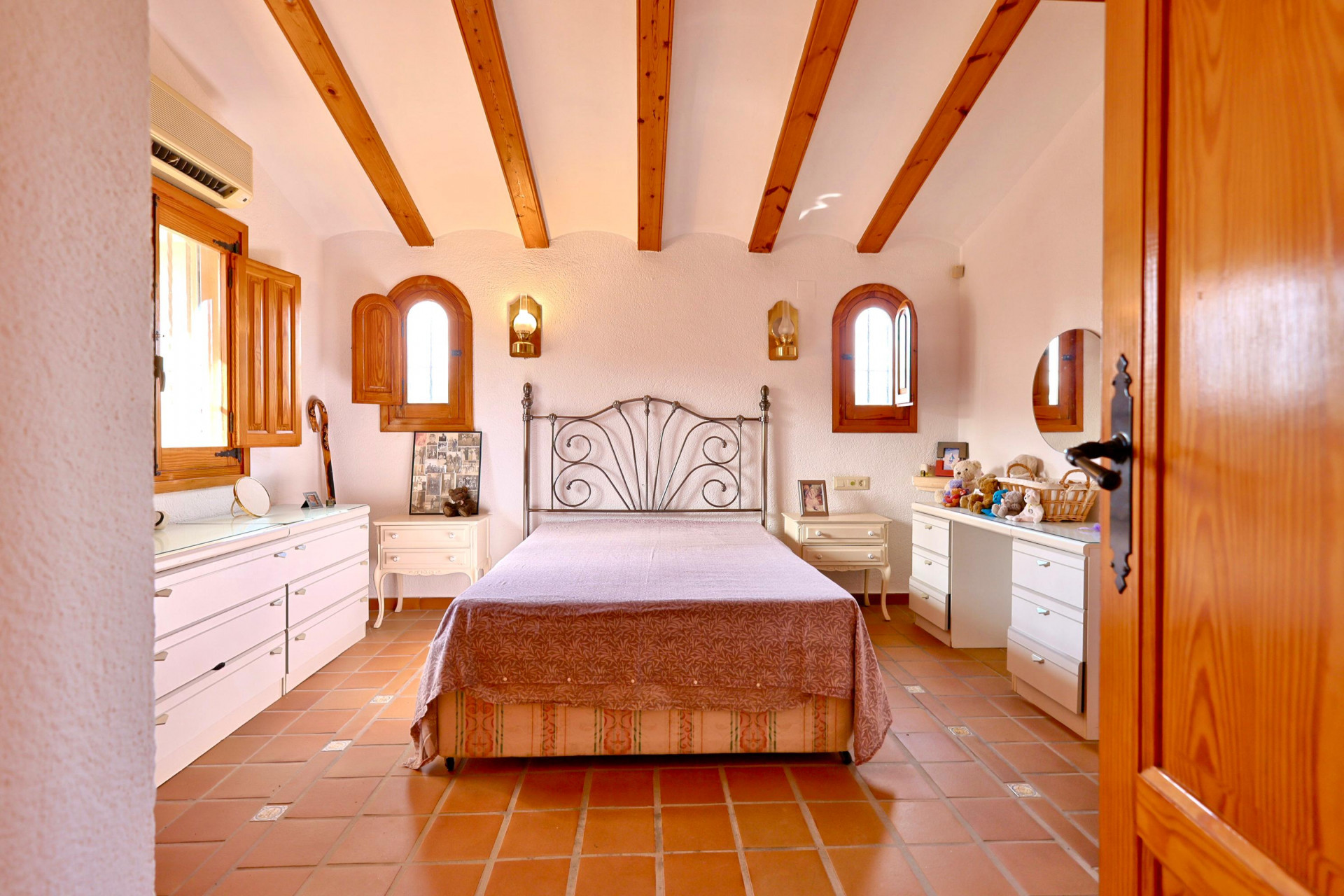 Dormitorio rústico con vigas de madera a la vista, suelos de terracota y ventanas de arco con contraventanas de madera. Incluye mobiliario blanco y cabecero de forja.
