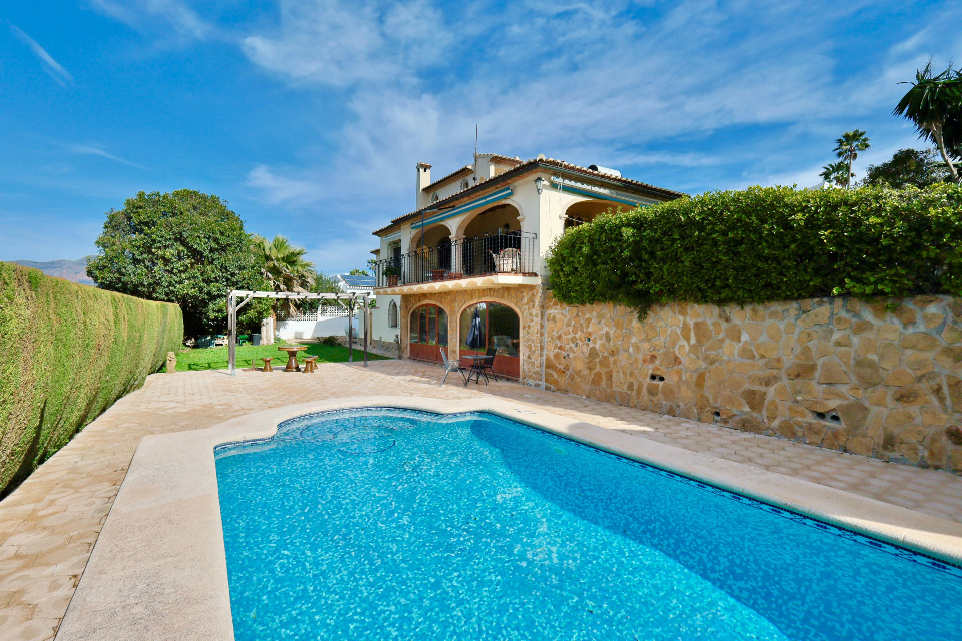 Exterior de villa mediterránea con piscina privada, amplia terraza pavimentada y fachada de piedra natural. Planta superior con arcos y barandilla de hierro forjado.