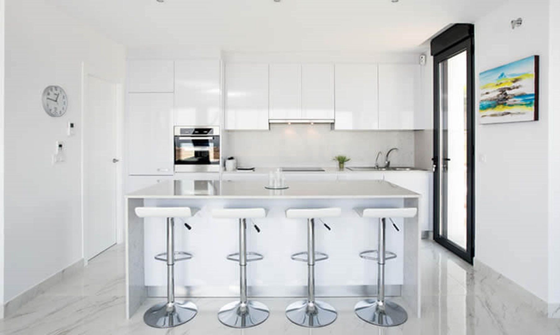 Cocina moderna de concepto abierto con mobiliario blanco brillante, isla central con encimera de cuarzo y cuatro taburetes. Suelos de gres porcelánico con efecto mármol.
