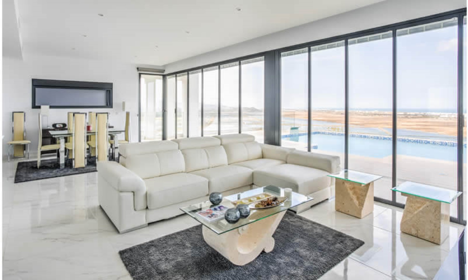 Salón comedor de planta abierta con suelos de mármol, grandes ventanales de suelo a techo y vistas directas a la piscina infinita y el paisaje mediterráneo.