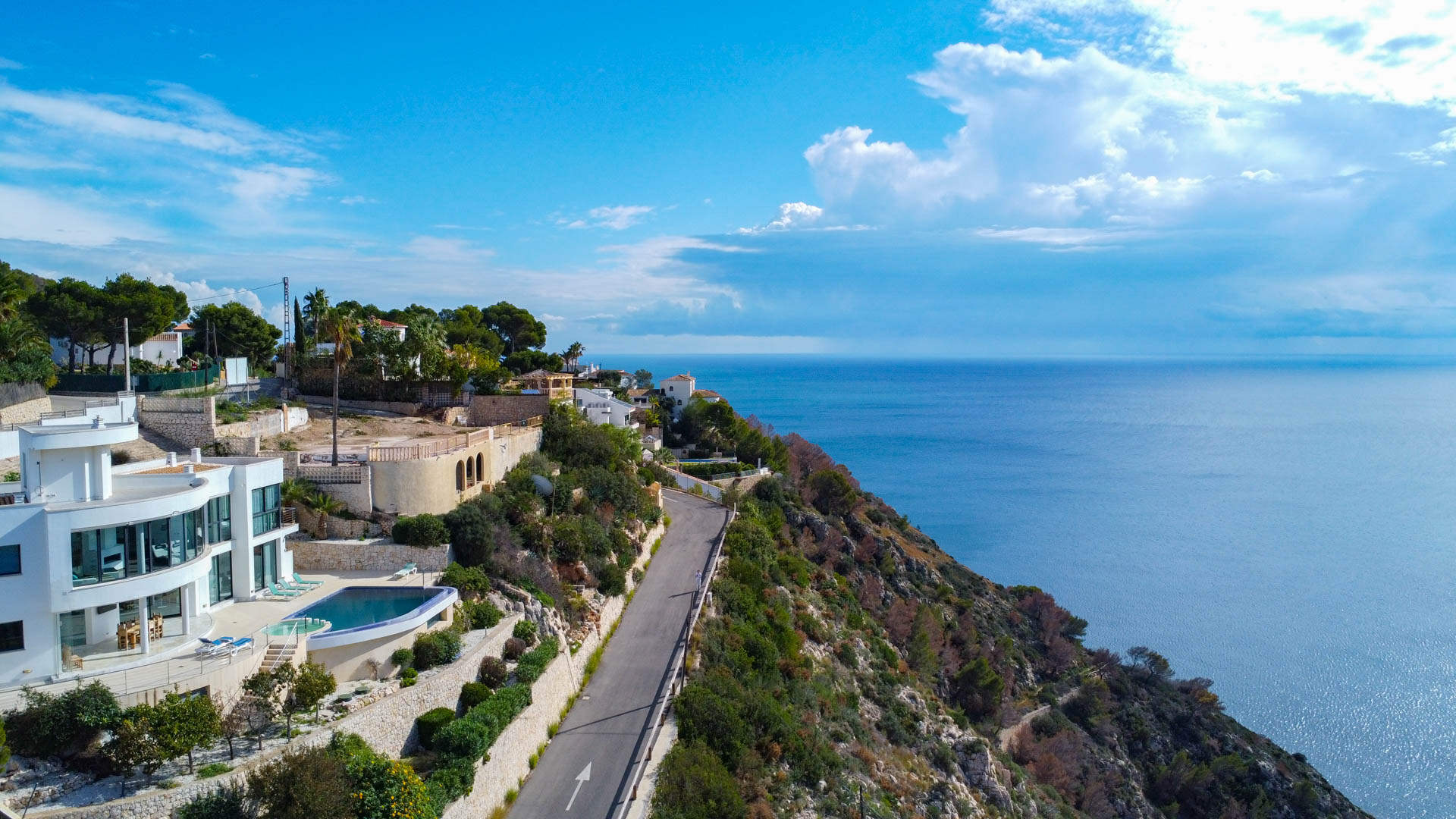 Vista aérea de la villa moderna con fachada curva, piscina infinita y amplios ventanales, situada en un acantilado con vistas panorámicas al mar Mediterráneo en Jávea.