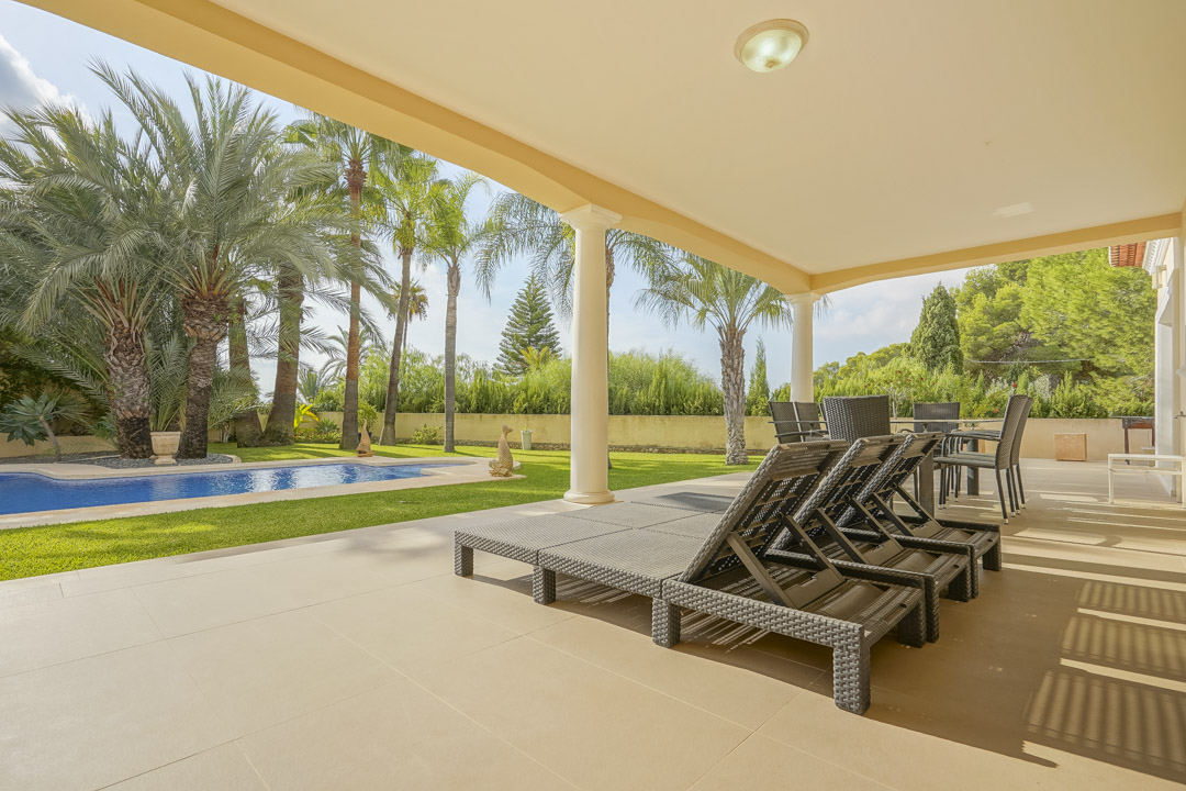 Amplia terraza cubierta con columnas clásicas y mobiliario exterior, con vistas directas a la piscina y al jardín mediterráneo con palmeras.