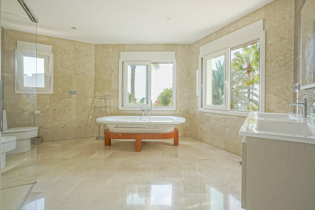 Amplio baño principal revestido en mármol con bañera exenta sobre base de madera, lavabo doble y vistas despejadas a las palmeras del jardín.