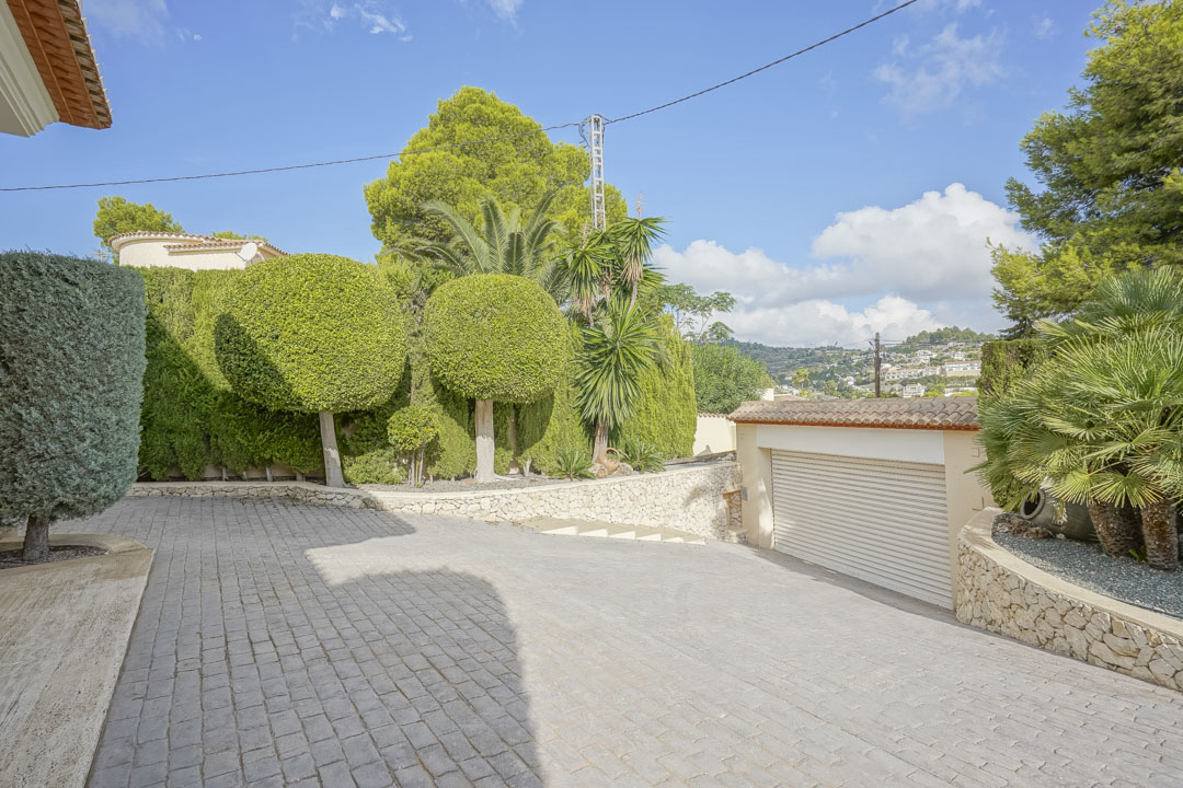 Amplio camino de entrada pavimentado con acceso al garaje, rodeado de un jardín mediterráneo maduro con árboles ornamentales y muros de piedra natural.
