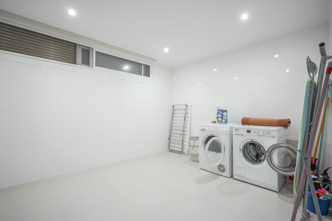Amplio lavadero situado en el sótano, equipado con lavadora, secadora y paredes alicatadas en blanco. Incluye iluminación empotrada y una ventana alta para ventilación.
