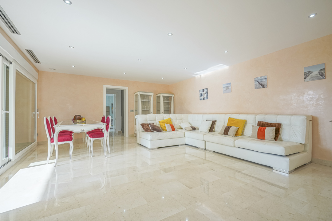 Amplio salón-comedor con suelos de mármol, un gran sofá rinconera de cuero blanco y acceso directo al exterior mediante ventanales correderos bajo iluminación empotrada.