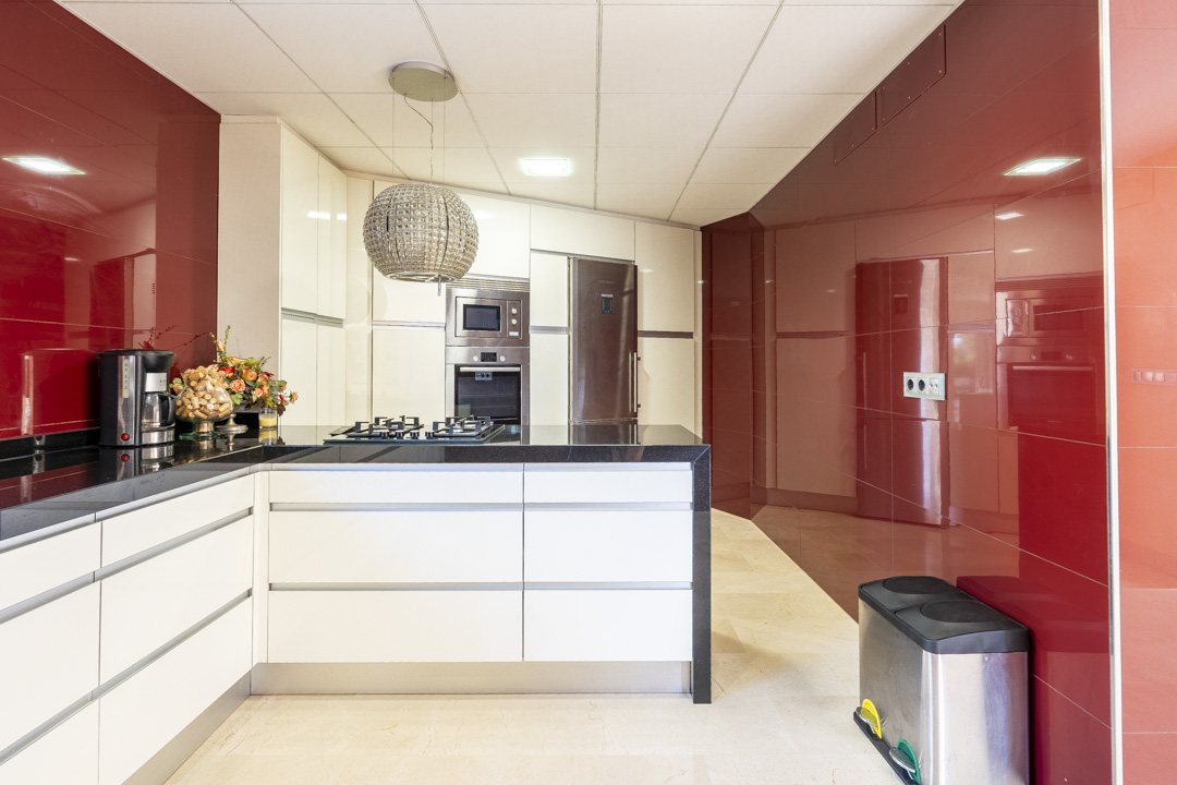 Cocina moderna con armarios blancos sin tiradores, encimera de granito negro y paredes de paneles rojos brillantes. Incluye electrodomésticos integrados y una lámpara colgante esférica.