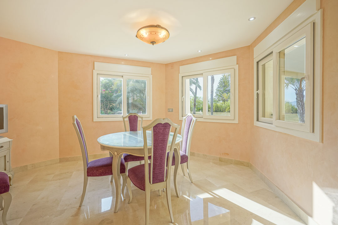 Comedor luminoso con mesa circular de cristal, sillas con tapicería violeta, suelo de mármol pulido y vistas al jardín mediterráneo a través de ventanas correderas.