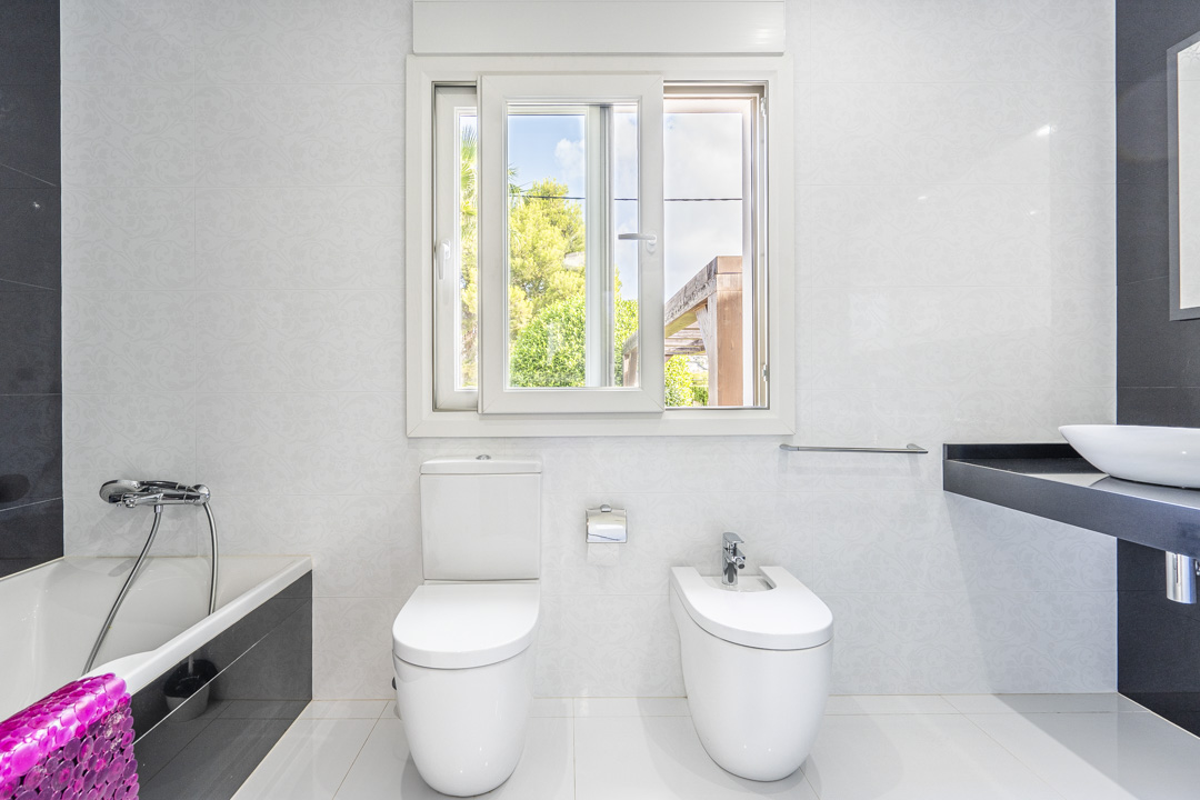 Cuarto de baño moderno con inodoro, bidé y bañera. Incluye un lavabo sobre encimera negra, azulejos blancos con relieve y una ventana corredera con vistas al jardín.