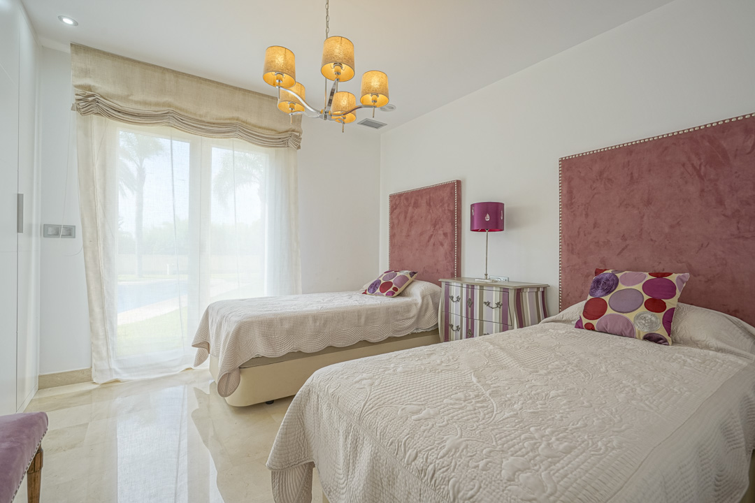 Dormitorio con dos camas individuales, cabeceros de terciopelo rosa y acceso directo a la terraza. Presenta suelos de mármol, iluminación empotrada y una lámpara de araña clásica.