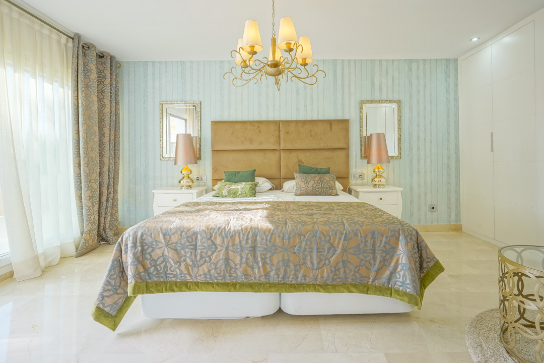 Dormitorio elegante con papel pintado turquesa, cabecero de terciopelo mostaza y suelos de mármol. Dispone de armarios empotrados blancos y gran entrada de luz natural.