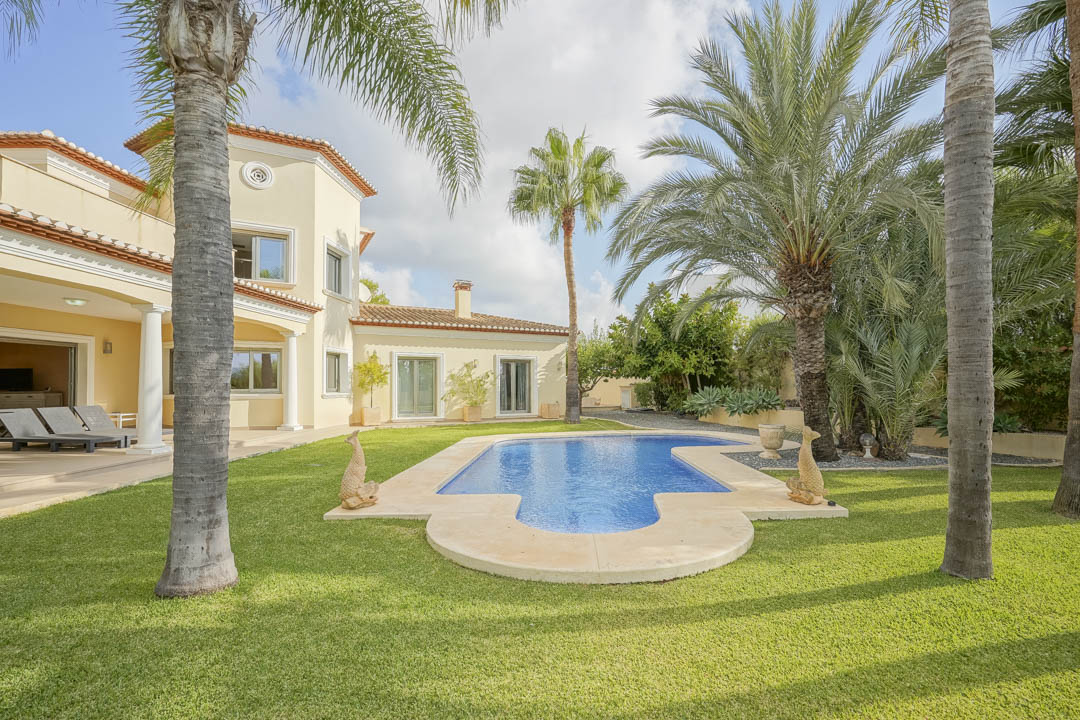 Piscina de forma irregular rodeada de césped natural y palmeras maduras frente a la fachada de estilo mediterráneo con terraza cubierta y columnas blancas.