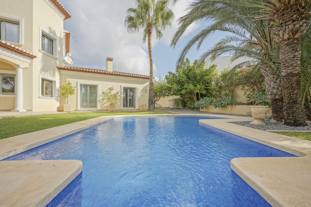 Piscina exterior rodeada de césped y un jardín mediterráneo con palmeras, junto a la fachada de color crema con carpintería verde y porches de estilo tradicional.