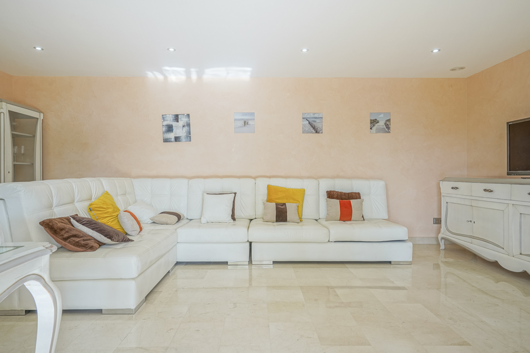 Salón con sofá rinconera de cuero blanco, suelos de mármol pulido y paredes en tono melocotón con iluminación empotrada y mobiliario clásico.