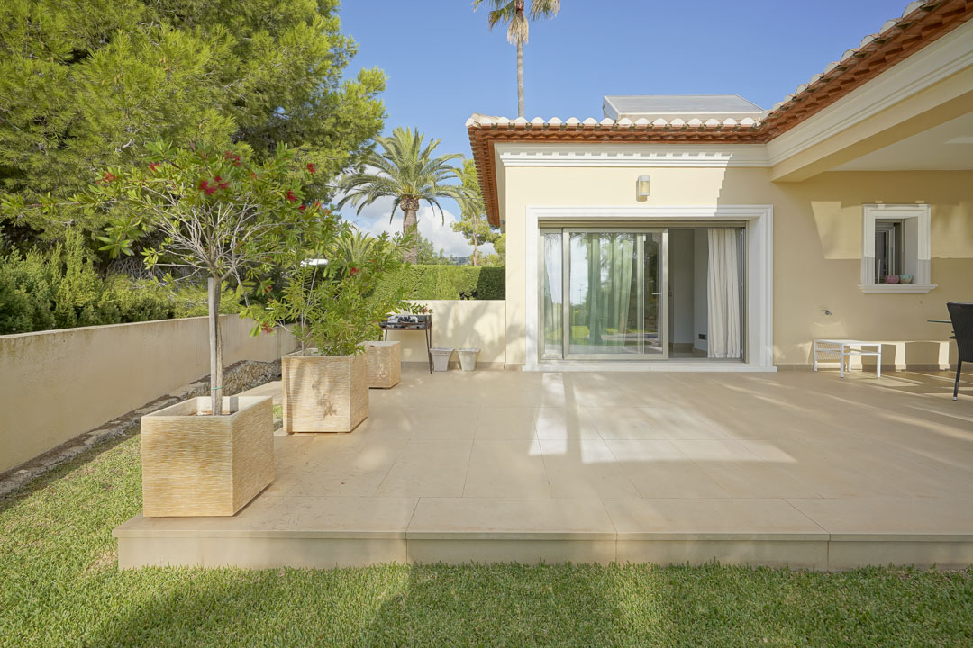 Terraza exterior con baldosas de tonos claros, acceso directo al interior por puerta corredera y jardín con pinos, palmeras y paneles solares en el tejado.