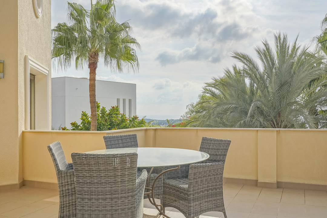 Terraza exterior con mesa de cristal y sillas de mimbre gris, rodeada de palmeras y vistas despejadas hacia el mar Mediterráneo.