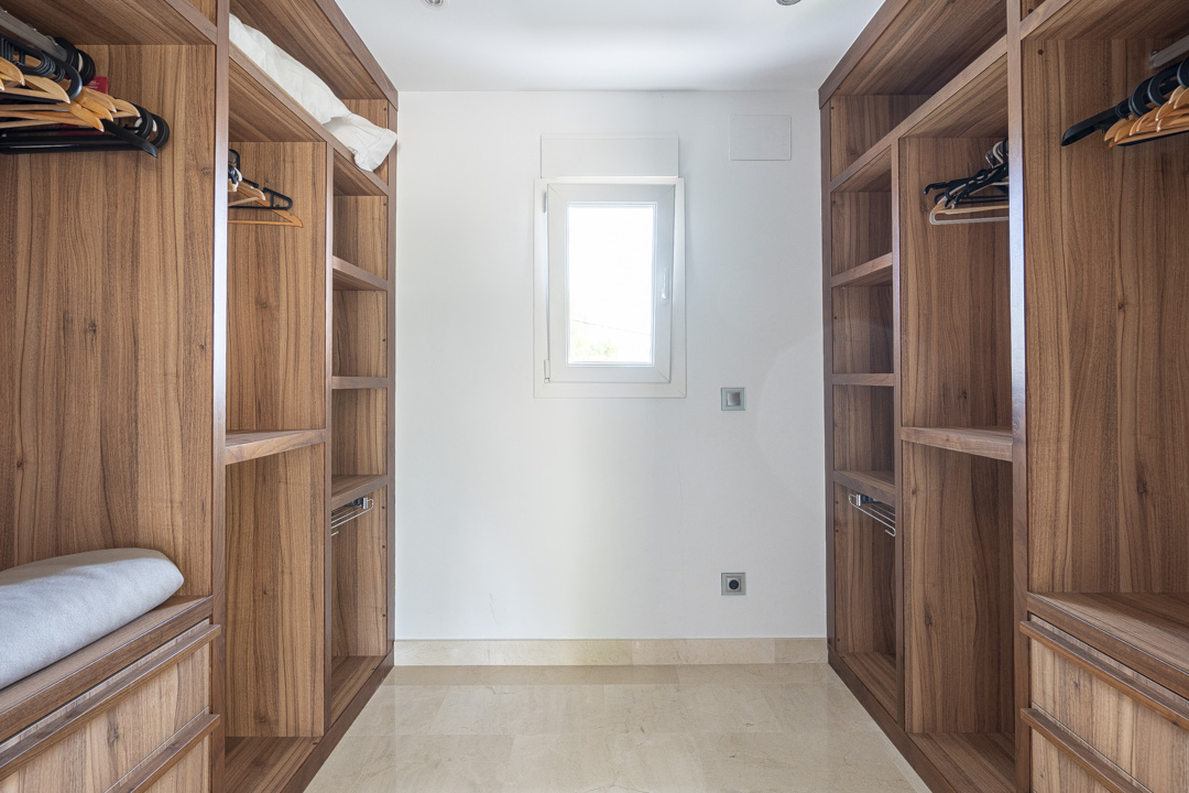 Vestidor con armarios de madera de nogal a medida, estanterías abiertas y cajoneras. Incluye una ventana central para luz natural y suelos de mármol pulido.