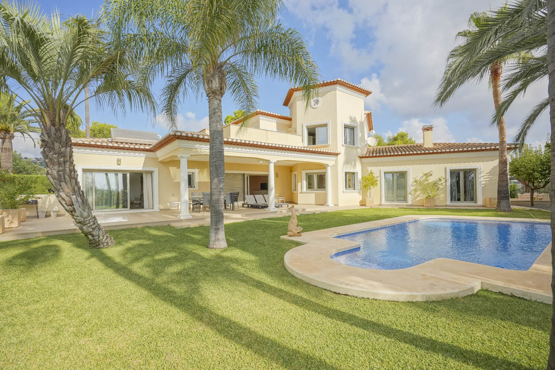 Vista exterior de la villa con piscina de forma irregular, rodeada de césped y palmeras maduras. Destaca la terraza cubierta con columnas blancas y la arquitectura tradicional.