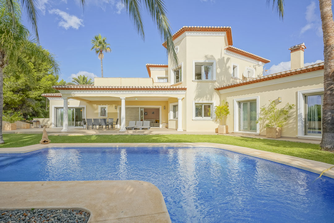 Vista exterior de la villa con piscina de gresite azul, porche cubierto con columnas blancas y jardín mediterráneo con palmeras maduras.
