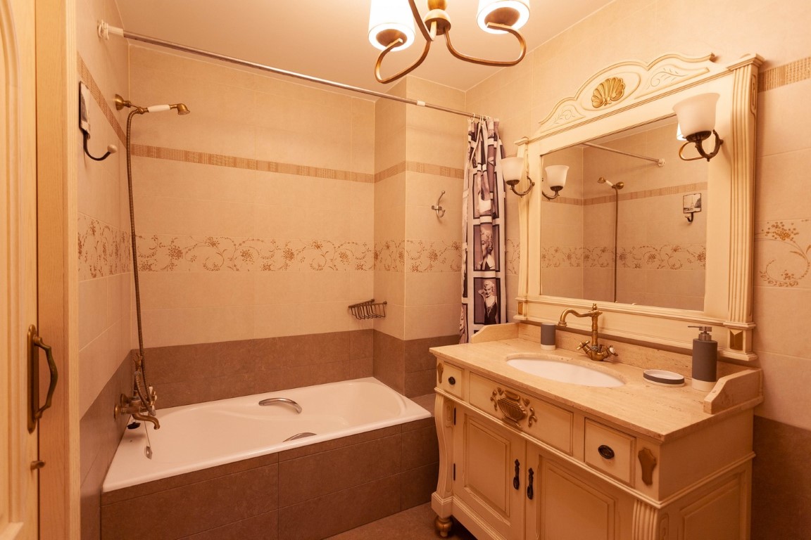 Baño de estilo clásico con tocador de madera tallada, encimera de piedra y grifería de bronce. Incluye bañera integrada y azulejos decorativos en tonos crema.