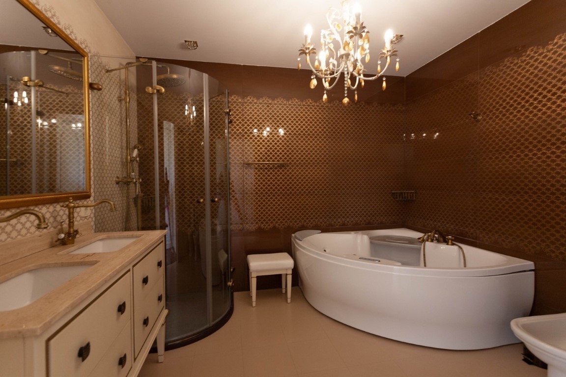 Baño principal con bañera de hidromasaje, ducha de cristal curva y lavabo doble con encimera de piedra. Paredes decoradas con azulejos marrones texturizados y grifería de latón.