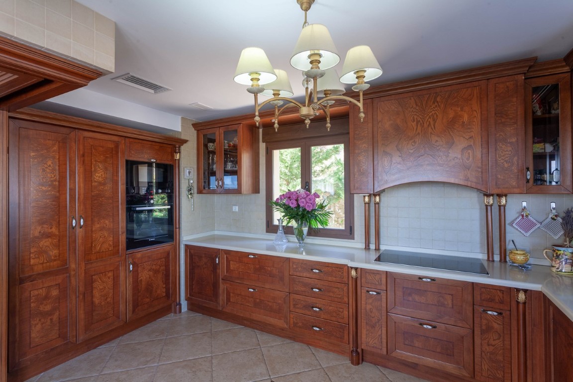 Cocina de diseño clásico con armarios de madera veteada, encimera clara y electrodomésticos integrados. Dispone de placa de inducción y una lámpara de araña central.