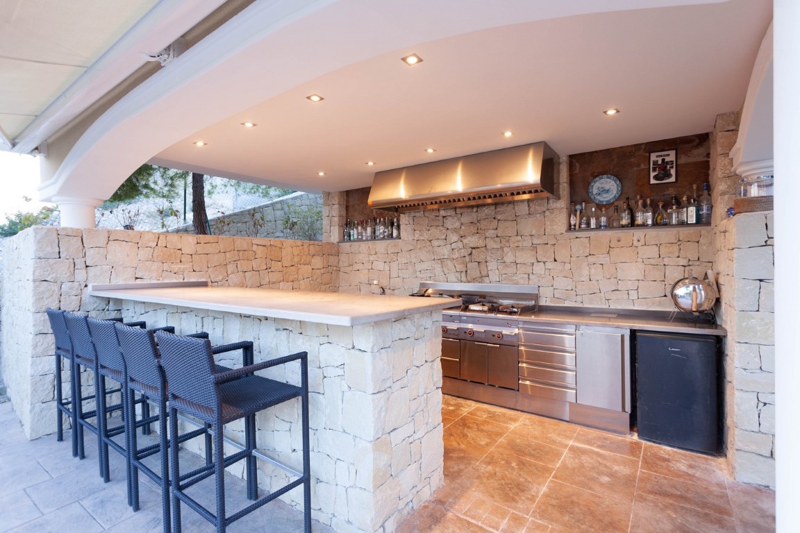 Cocina exterior profesional con paredes de piedra natural, encimera de piedra, electrodomésticos de acero inoxidable y barra con taburetes de mimbre bajo iluminación empotrada.