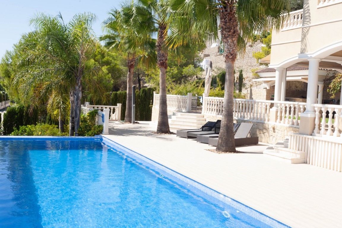 Piscina exterior con azulejos azules rodeada de una terraza de piedra clara, palmeras altas y una zona de descanso con tumbonas frente a la fachada de la villa.