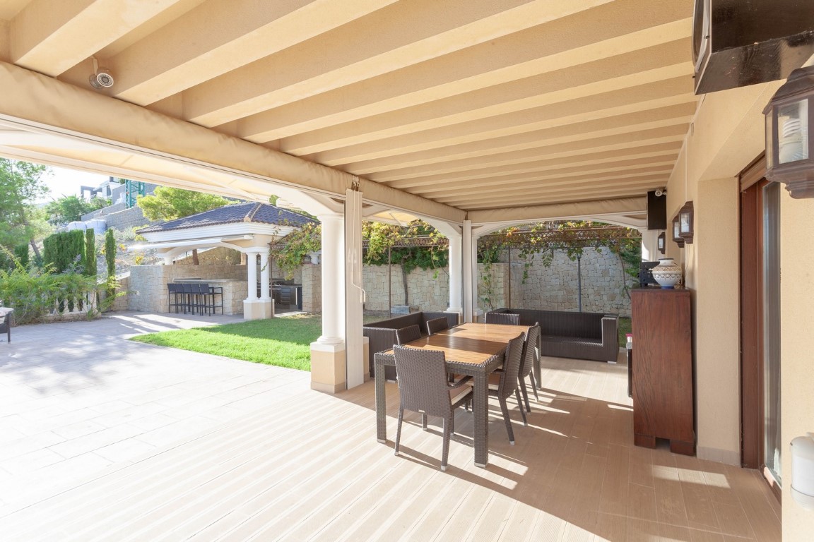 Terraza cubierta con techo de vigas, zona de comedor exterior y sofás de mimbre, con vistas a la cocina de verano y al jardín mediterráneo.