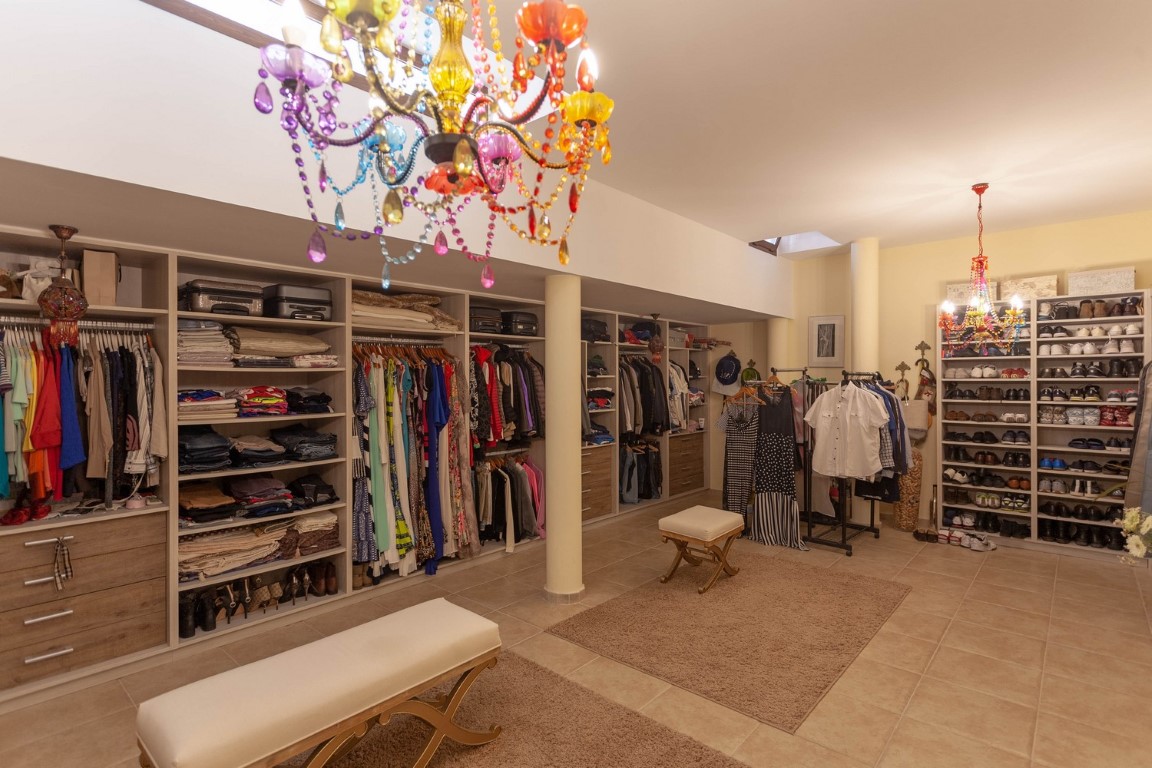 Vestidor diáfano con estanterías abiertas, cajoneras de madera y lámparas de araña multicolor. Dispone de bancos tapizados, claraboyas y una amplia zona de zapatero.