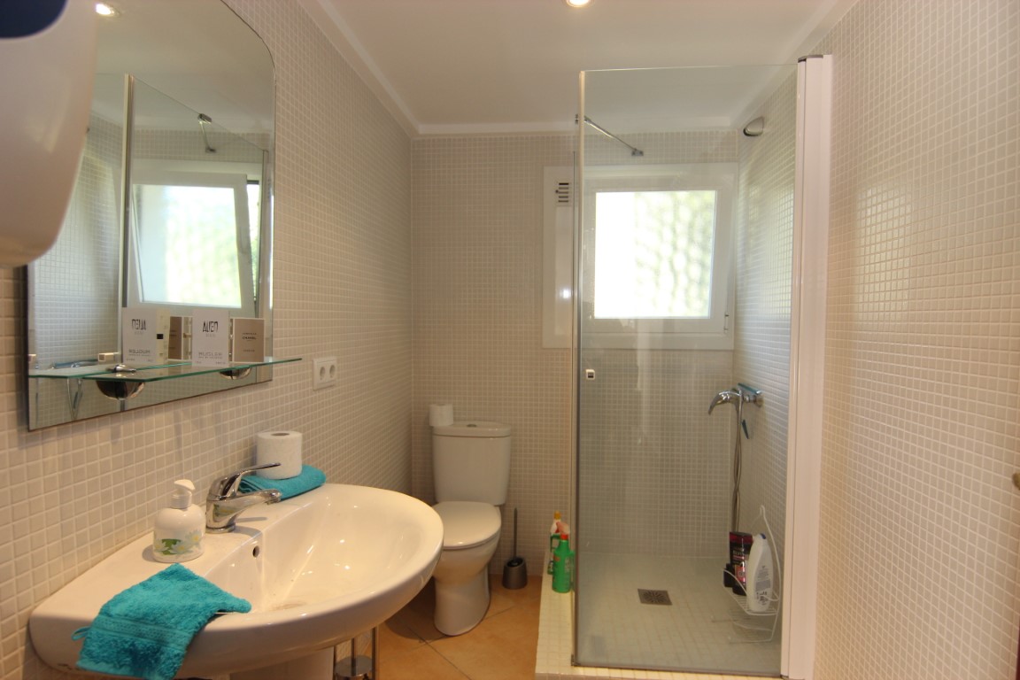 Baño funcional con paredes revestidas en mosaico beige, ducha con mampara de cristal y lavabo cerámico blanco. Incluye una ventana para ventilación y luz natural.