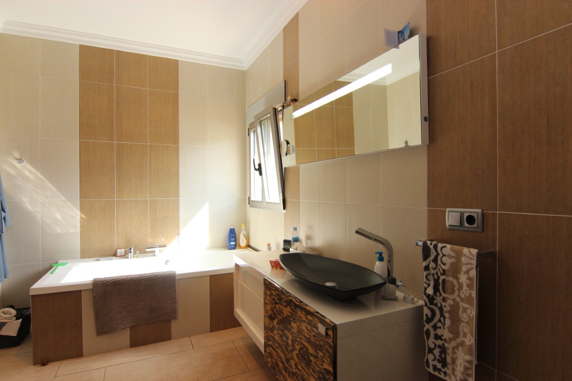 Baño moderno con bañera integrada, lavabo de diseño sobre encimera y paredes alicatadas en tonos crema y madera. Incluye un gran espejo con iluminación integrada y luz natural.