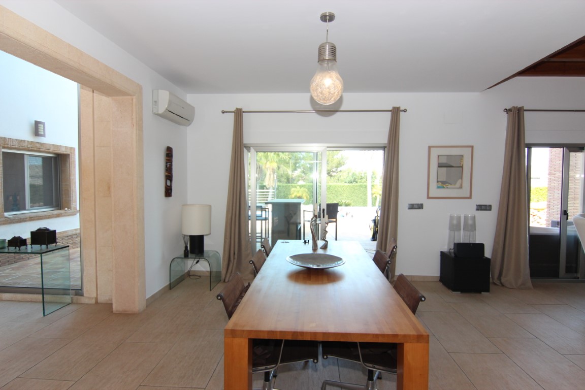 Comedor con mesa de madera maciza y suelos cerámicos, conectado a la terraza exterior mediante amplios ventanales que aportan abundante luz natural.