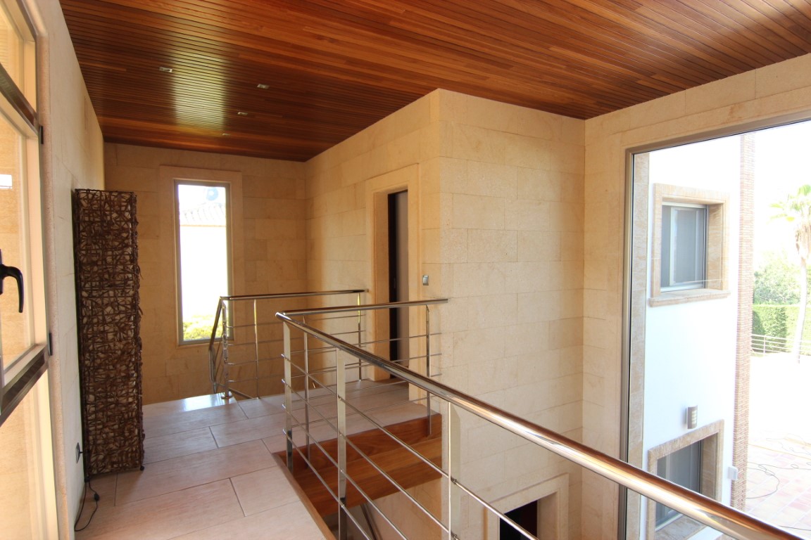 Distribuidor de la planta superior con paredes de piedra natural, techos de madera y barandillas de acero inoxidable sobre el hall de doble altura.
