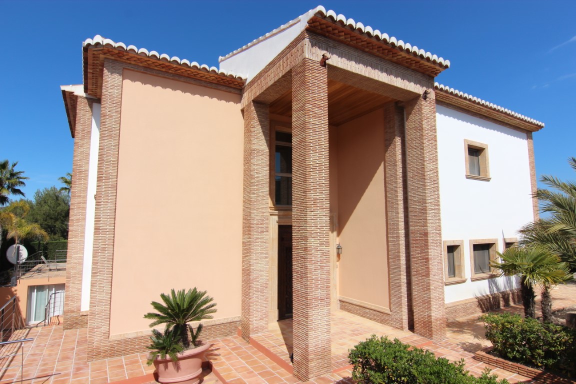 Fachada principal con entrada de doble altura, destacando las columnas de ladrillo visto, techos de madera y suelos de terracota rodeados de vegetación mediterránea.