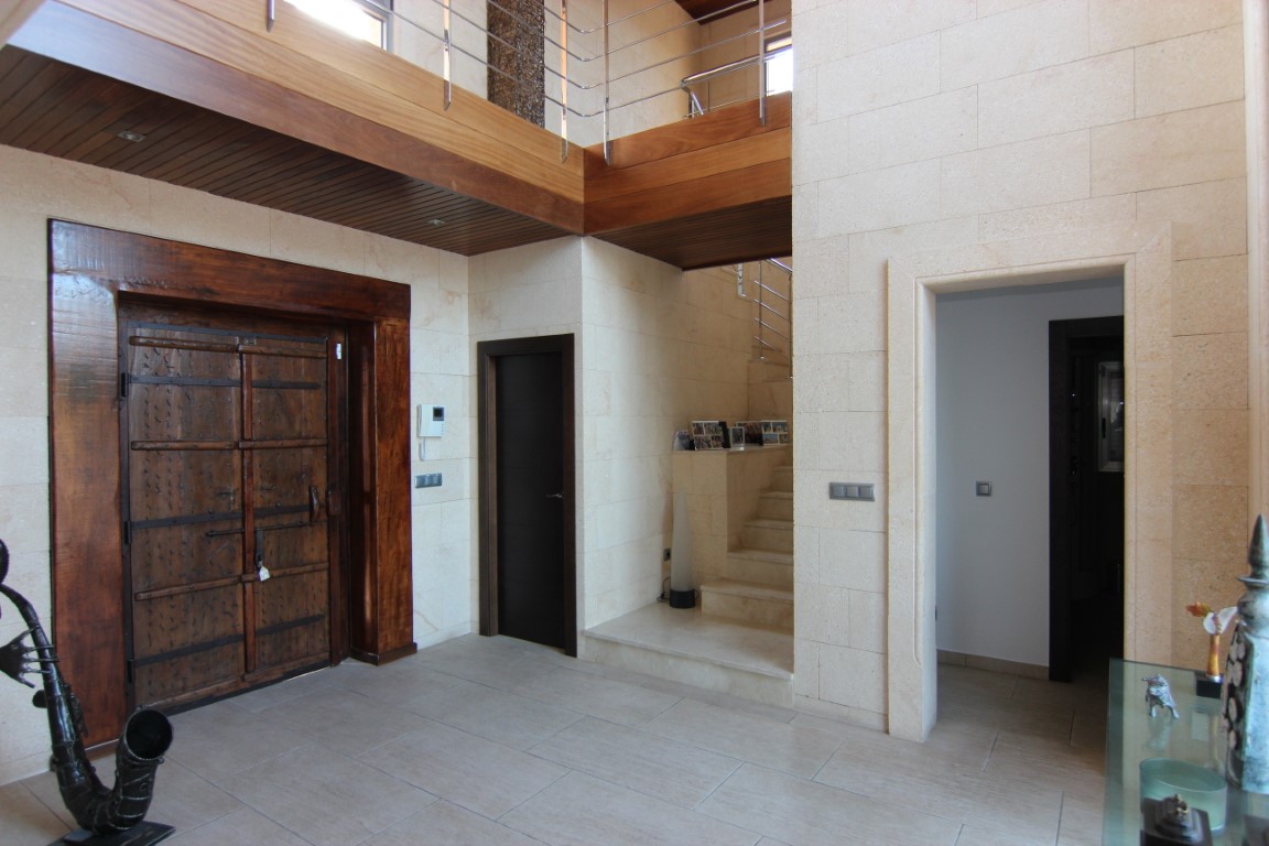 Hall de entrada de doble altura con paredes de piedra natural, una gran puerta rústica de madera y una galería superior con techos panelados en madera.