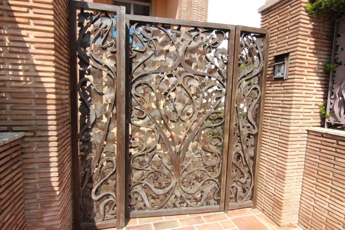 Puerta de entrada de metal artesanal con diseño orgánico, integrada en pilares de ladrillo visto de estilo mediterráneo.