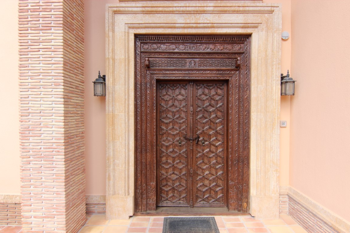 Puerta principal de madera tallada a mano con intrincados motivos geométricos, enmarcada en piedra natural y flanqueada por faroles clásicos y columnas de ladrillo visto.