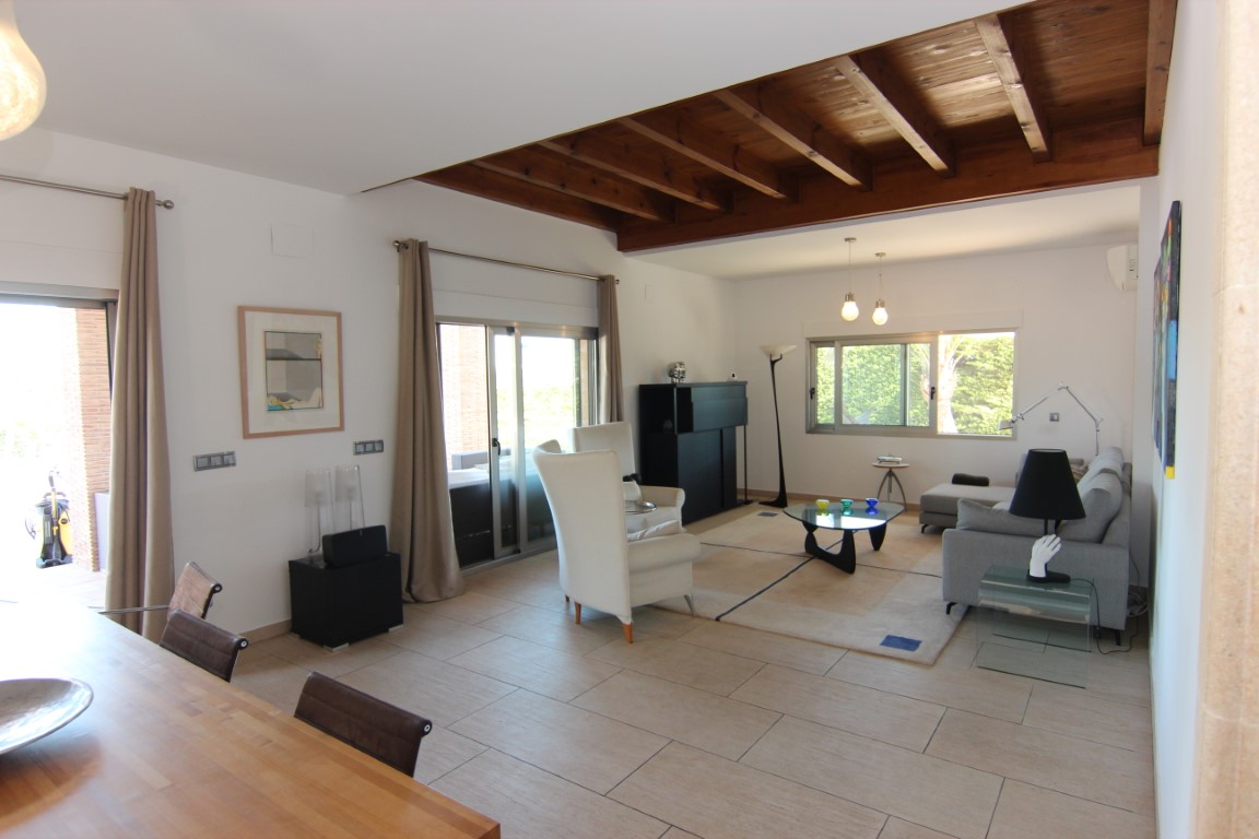 Salón luminoso con vigas de madera vista, suelos cerámicos claros y acceso directo a la terraza mediante amplios ventanales correderos.