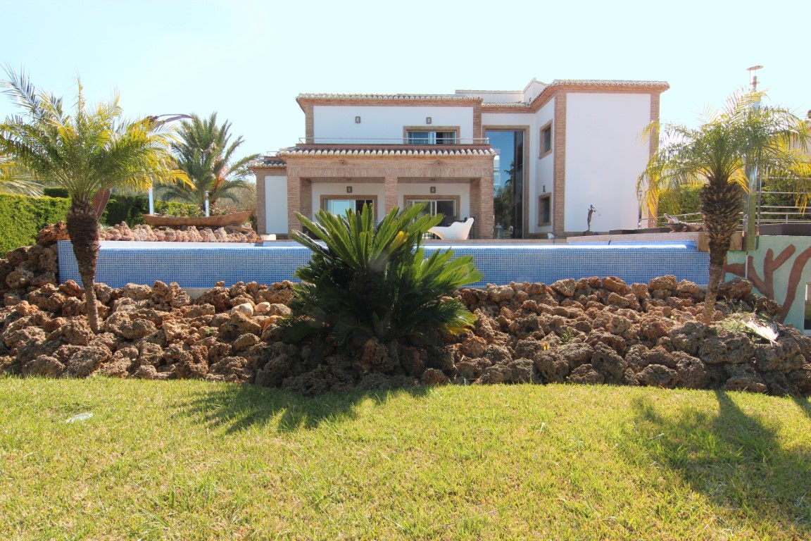 Vista exterior de la villa con piscina de mosaico azul, rodeada de jardines con roca volcánica y palmeras, destacando la fachada con detalles en ladrillo visto.