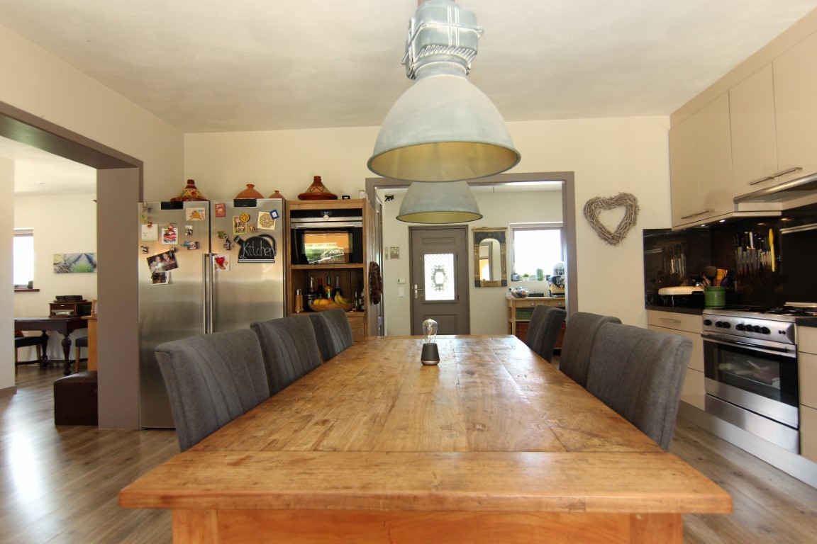 Cocina con comedor integrado, destacando una gran mesa de madera rústica, lámparas industriales y electrodomésticos de acero inoxidable en un espacio abierto.