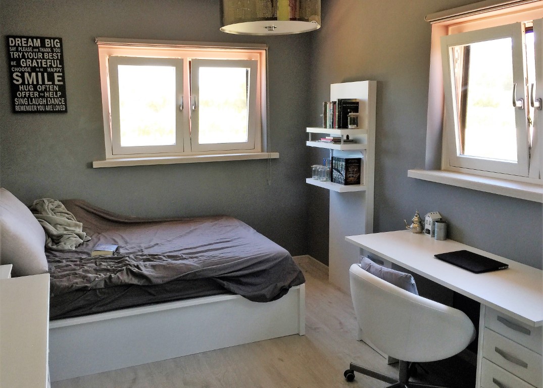 Dormitorio doble con zona de despacho, paredes en tono gris, escritorio blanco y suelos de madera clara. Dos ventanas proporcionan abundante luz natural al espacio.