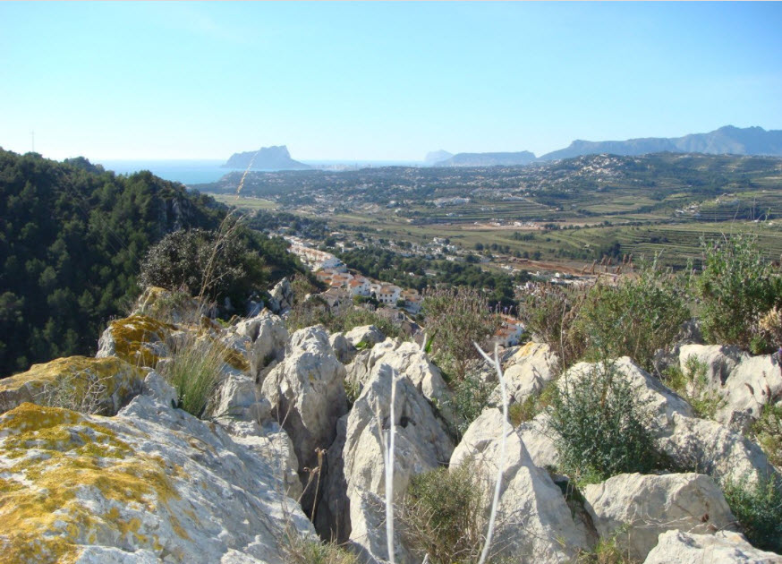 Vistas panorámicas desde el terreno elevado hacia el Peñón de Ifach y el mar Mediterráneo, rodeado de formaciones rocosas naturales y vegetación autóctona.